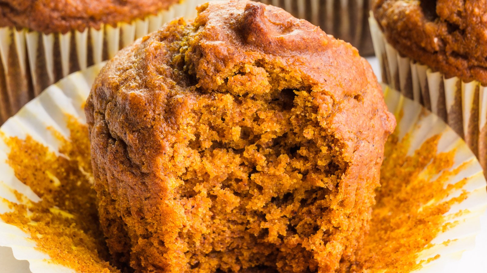 A vegan pumpkin muffin has a bite taken out, revealing a moist crumb inside. There are other muffins barely visible in the background.