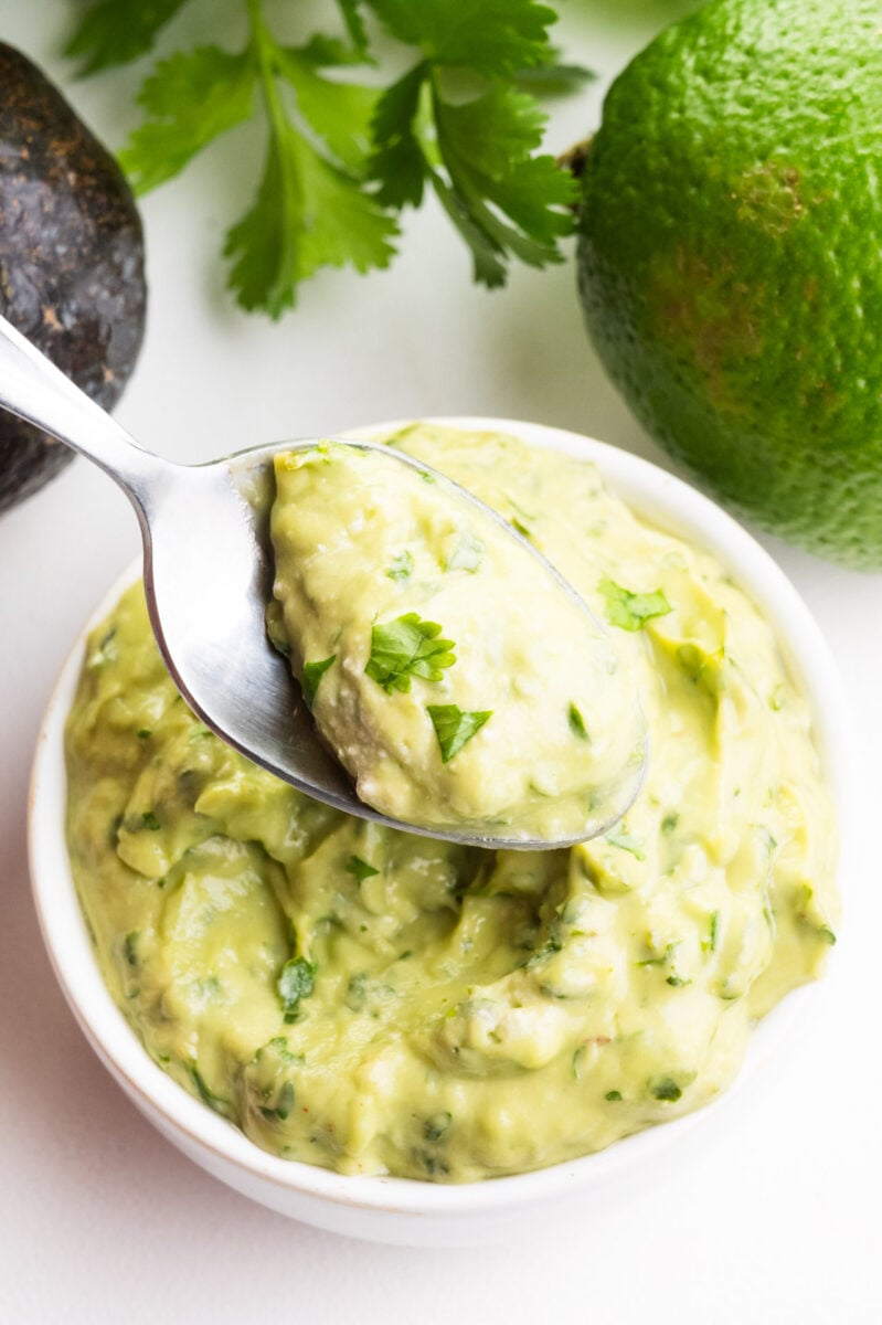 A spoonful of green avocado sauce hovers over a bowl of the sauce. There is an avocado, fresh cilantro, and a lime in the background.