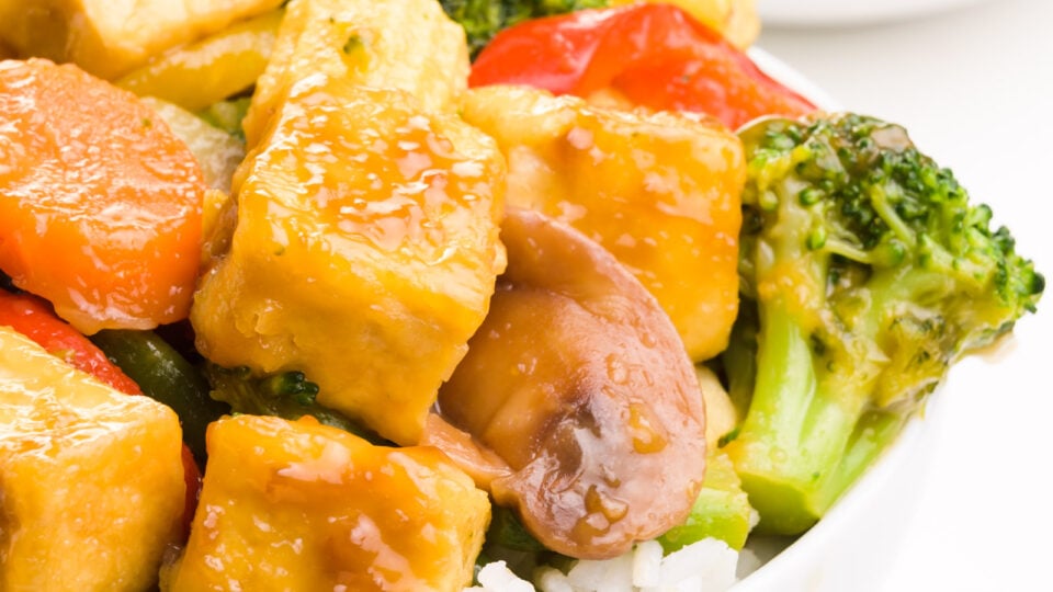 A closeup of glazed fried tofu in a plate over rice with steamed veggies.