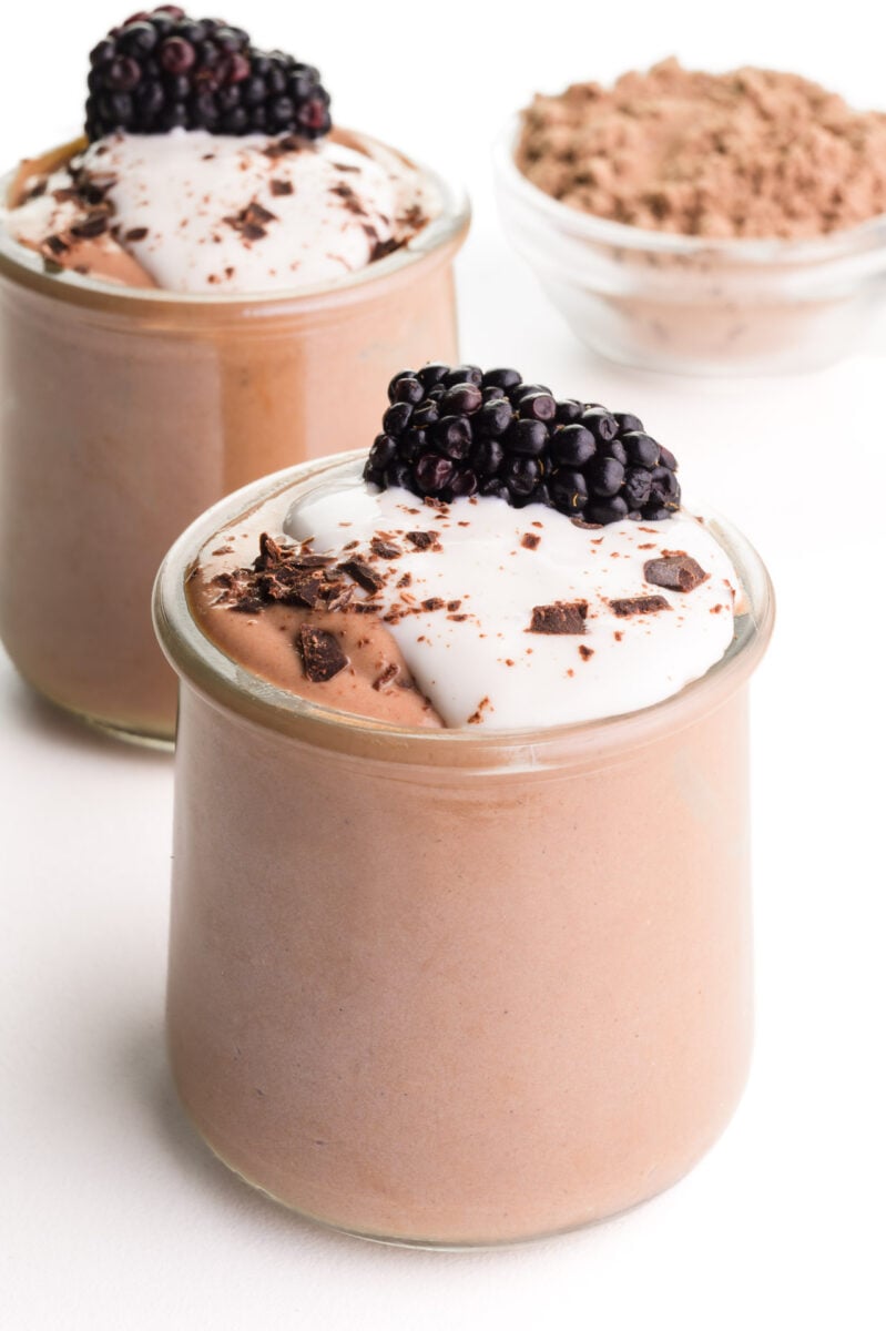 Two glass jars are full of protein pudding, topped with yogurt, chocolate shavings, and a blackberry. There is a bowl of chocolate protein powder in the background.
