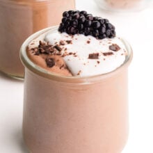 A closeup of a serving dish full of chocolate protein pudding, topped with yogurt, chocolate shavings, and a fresh blackberry.