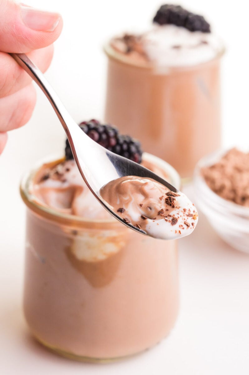 A hand holds a spoonful of protein pudding, hovering over two glass jars full of the pudding.