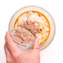 A hand pours chocolate protein powder into a bowl with other ingredients, like yogurt and vanilla extract.