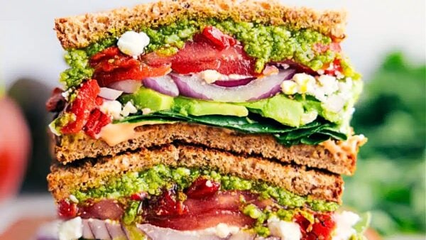 A sandwich is cut in half and stacked on the other half, revealing lots of veggies inside, like avocados, red onions, tomatoes, pesto, and more.