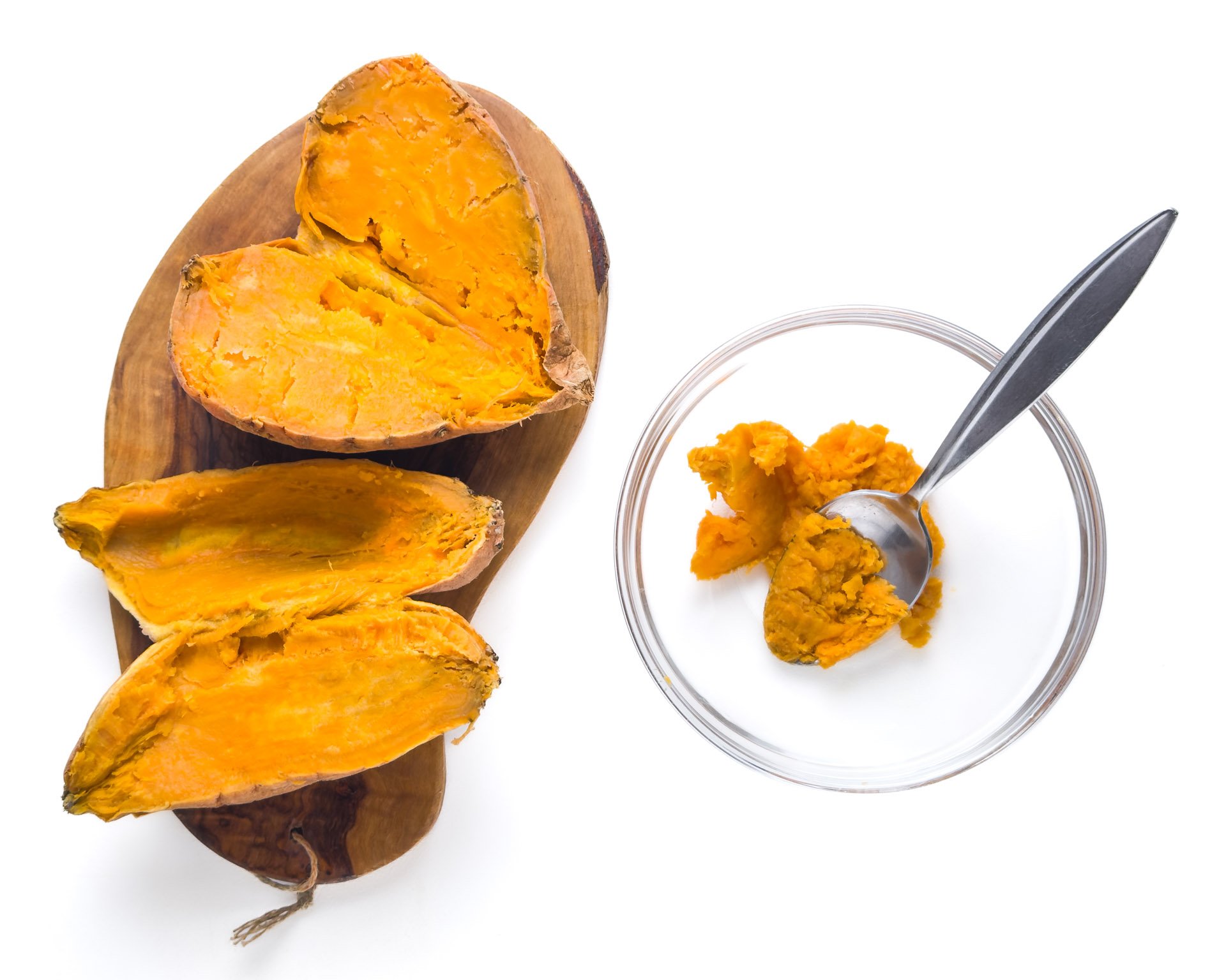 Looking down on cooked sweet potatoes on a cutting board. One has been partially scooped and the flesh is in a bowl with a spoon.