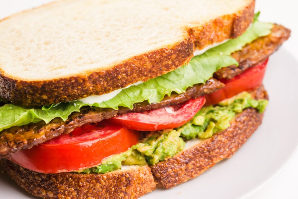 A vegan BLT sandwich on a plate shows layers of mashed avocado, tomatoes, vegan bacon, and lettuce.