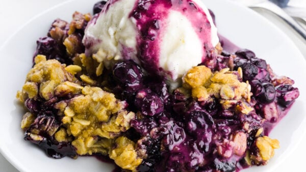 A blueberry crumble serving on a plate is topped with ice cream.