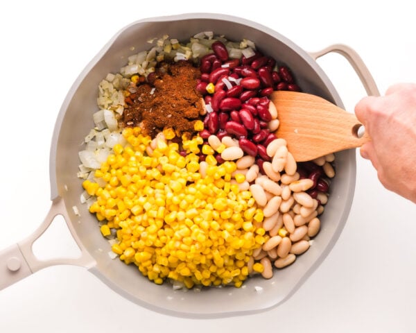 A hand holds a wooden spoon, stirring ingredients in a pan.