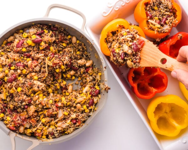 Quinoa mixture is being spooned from a skillet into a pan filled with bell pepper shells.