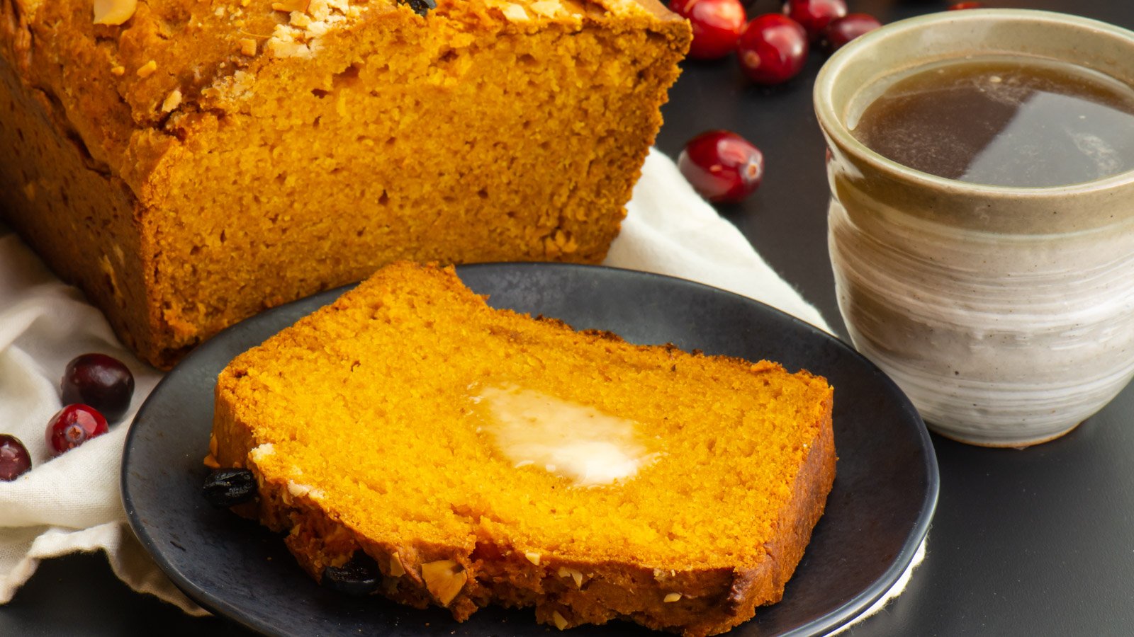A slice of pumpkin bread has a pat of melted butter on top. It sits in front of the rest of the loaf.
