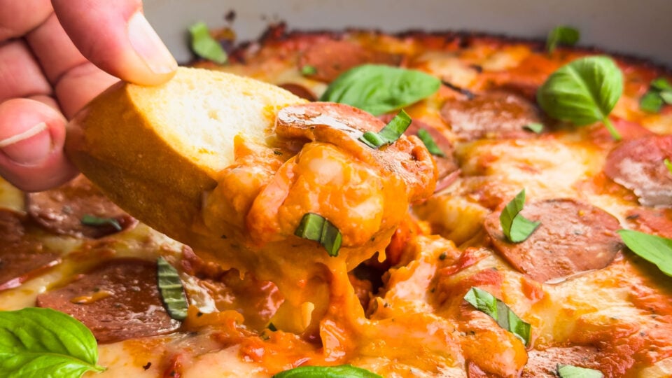A hand dips pizza dip from a skillet using a baguette slice.
