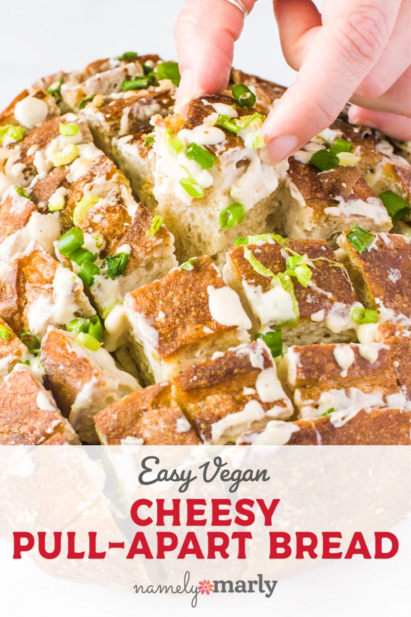 A hand is pulling a piece from a large loaf of bread that is covered in green onions and vegan cheese