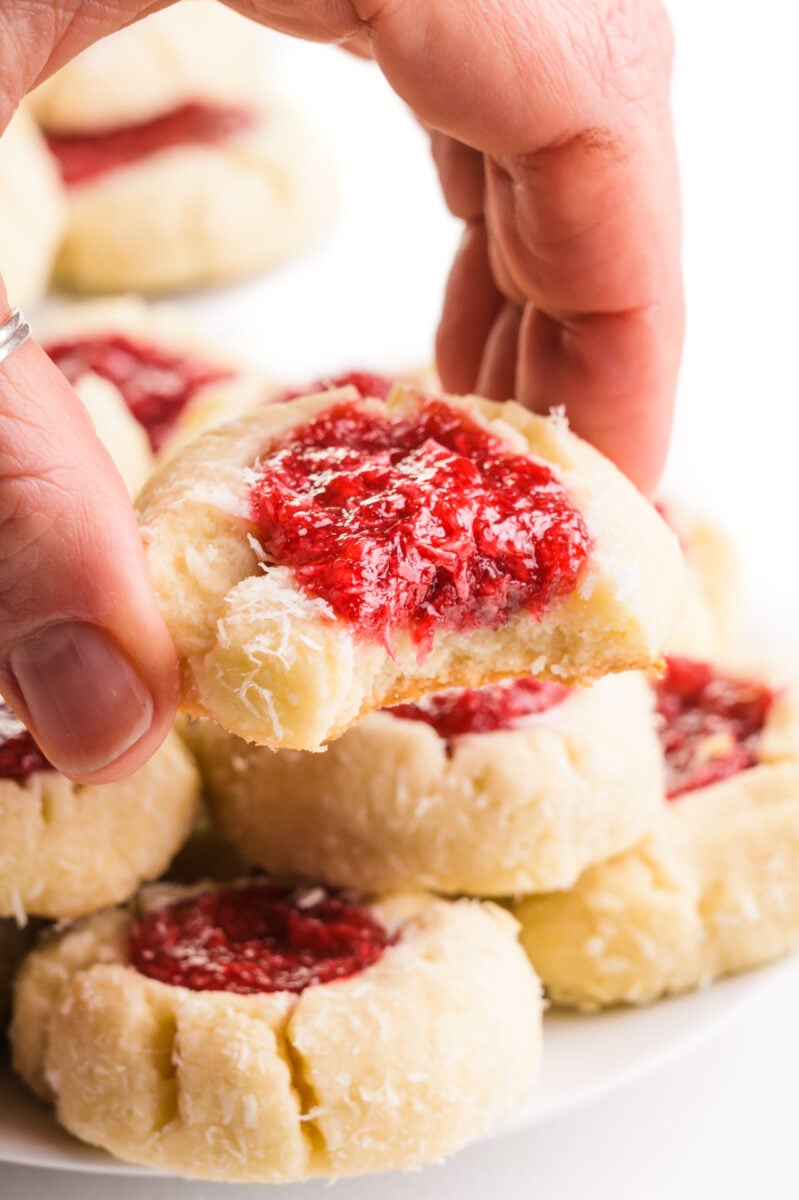 A hand holds a raspberry thumbprint cookie with a bite taken out.