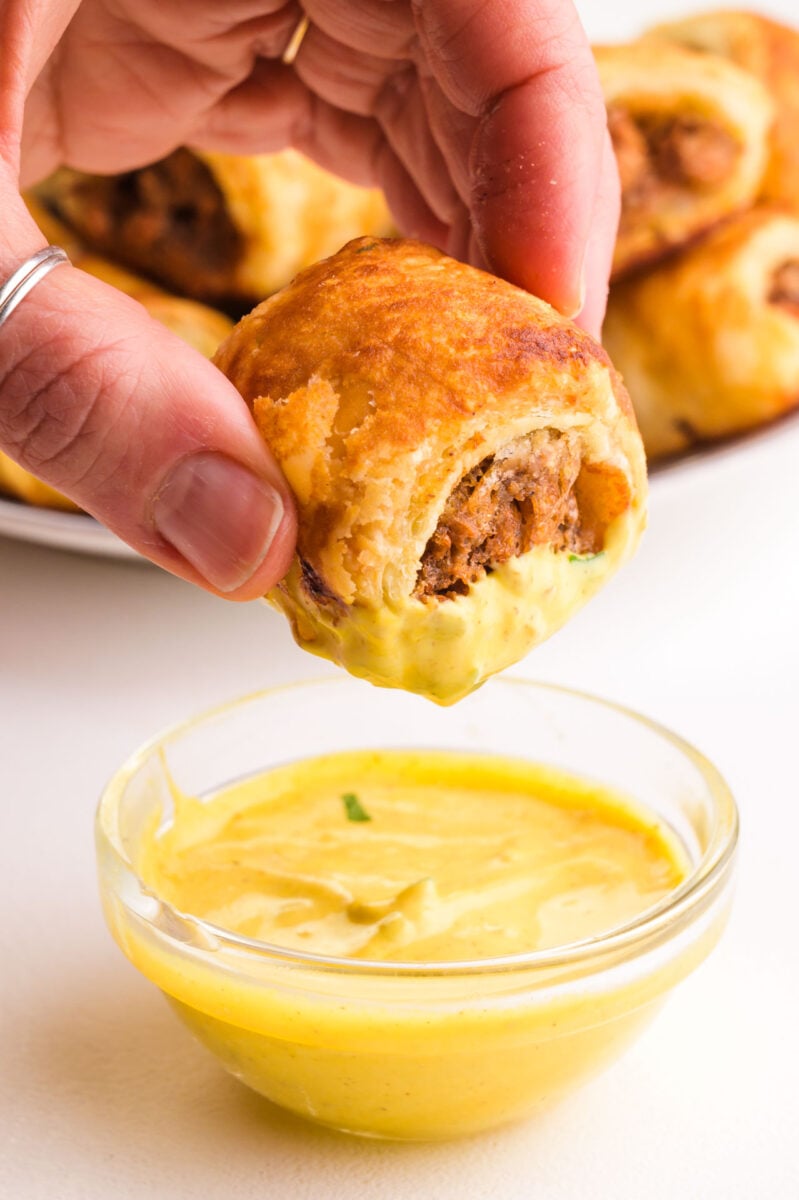 A hand dips a vegetarian sausage roll in a bowl of mustard sauce.
