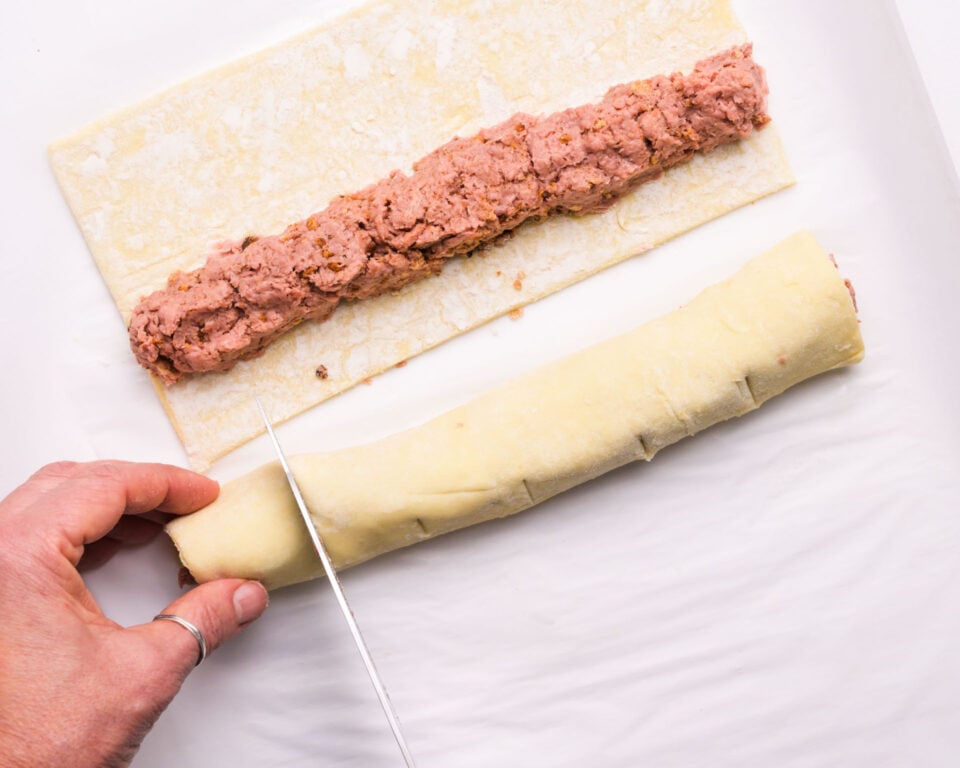 A hand cuts sausage rolls while the puff pastry sheet next to it has a roll of sausage on it.