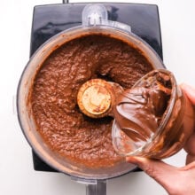 Melted chocolate is being poured into a food processor with chocolate brownie batter in it.