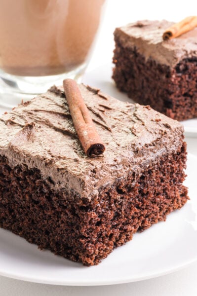 A slice of chocolate chai cake has frosting on top and a cinnamon stick. It sits in front of a cup of chai tea.