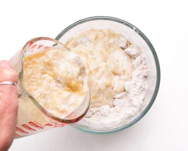 A mashed banana mixture is being poured into a bowl with a flour mixture.