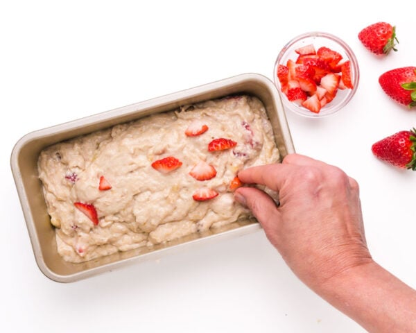 A hand places chopped strawberries on top of batter in a loaf pan.