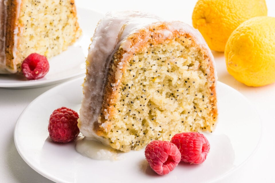 A slice of lemon cake on a plate has fresh raspberries next to it. There is another slice and fresh lemons in the background.