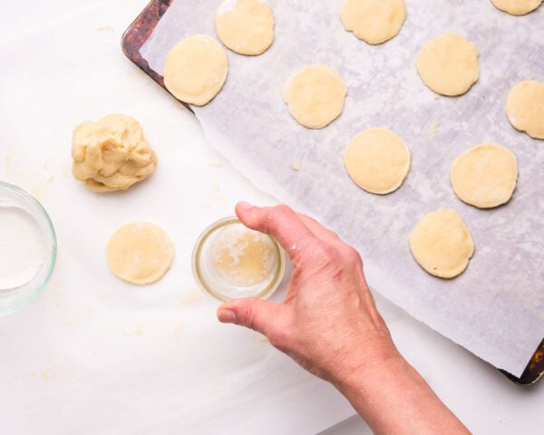 A hand presses a ball of dough under a glass to create a small circle of cookie dough for baking.