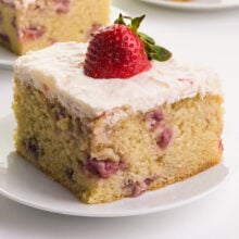 A closeup of a slice of strawberry cake on a plate with frosting on to and a fresh strawberry.