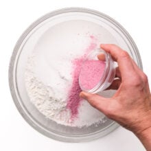 Strawberry jello mix is being poured into a mixing bowl with flour ingredients.