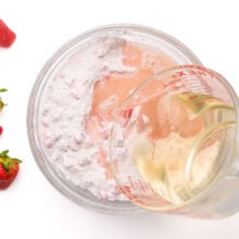 Wet ingredients are being poured into a mixing bowl. Fresh strawberries are by the bowl.