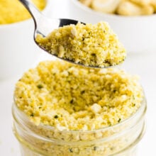 A hand holds a spoonful of cashew parmesan having over a glass jar full of it wit cashews behind it.