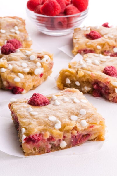Slices of raspberry white chocolate dessert bars on a table next to a bowl of fresh raspberries.