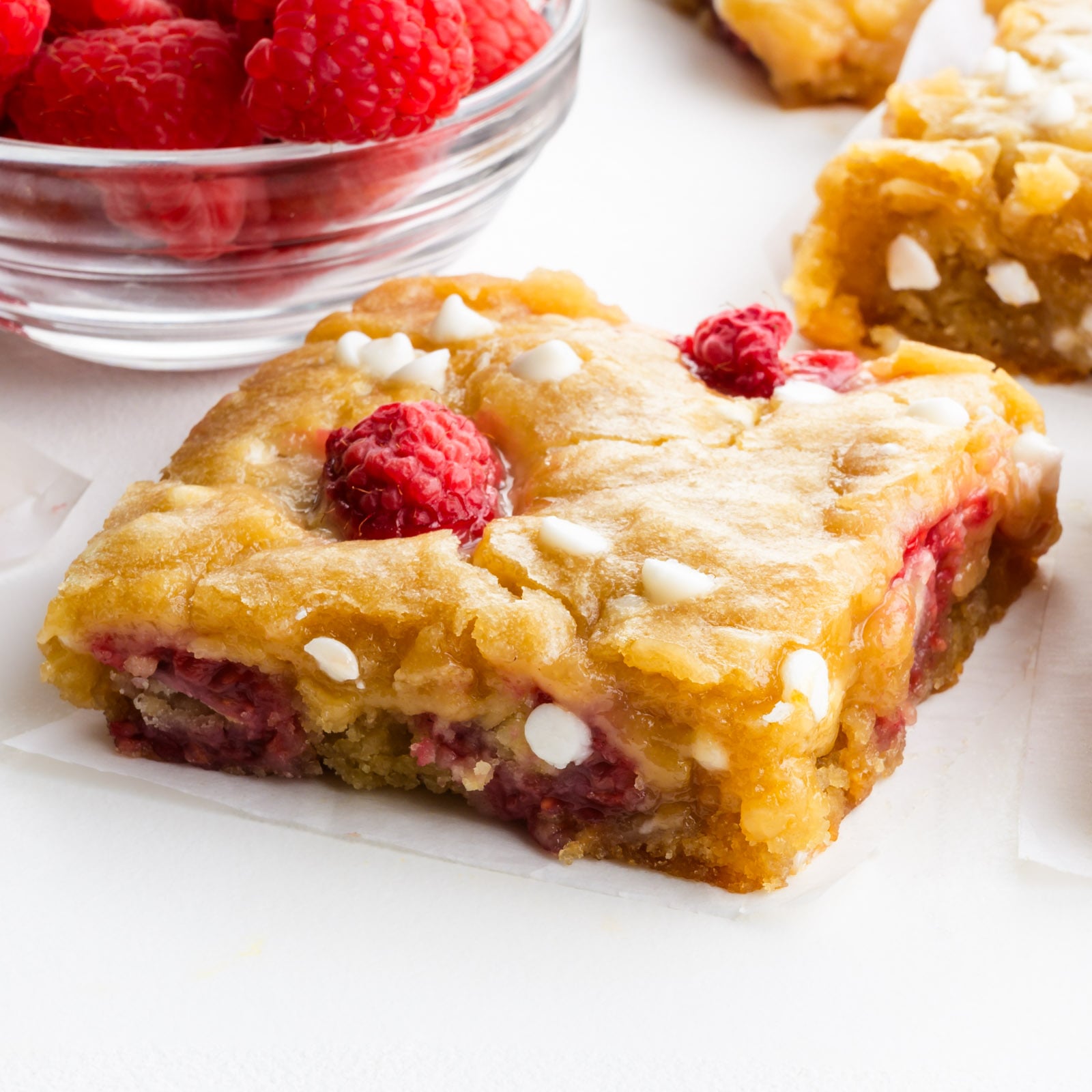 A raspberry white chocolate blondie slice on a table next to a bowl of fresh raspberries.