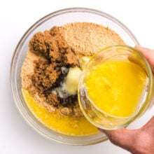 Melted butter is being poured into a bowl with graham cracker crumbs and other ingredients.