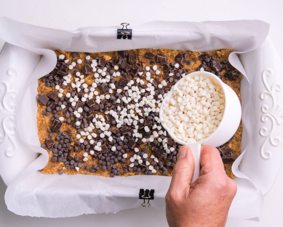 Sprinkling white chocolate chips on a layered dessert bar in a pan.