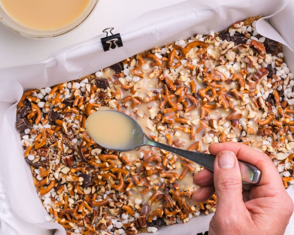 A hand drizzles sweetened condensed milk over a baking dish full of other ingredients.