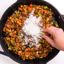 Flour is being sprinkled over veggies in an iron skillet.