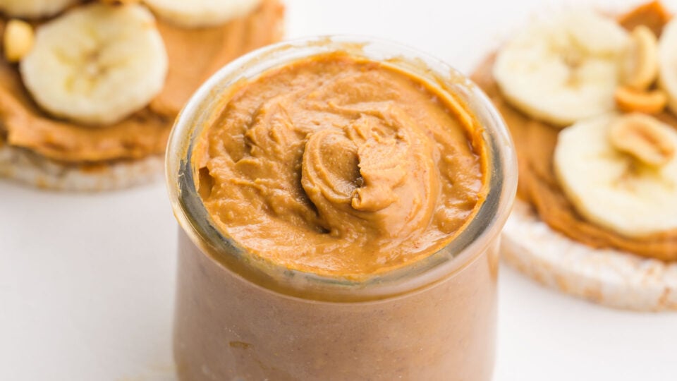 A closeup of a jar of peanut butter sitting in front of rice cakes with peanut butter and bananas on top.