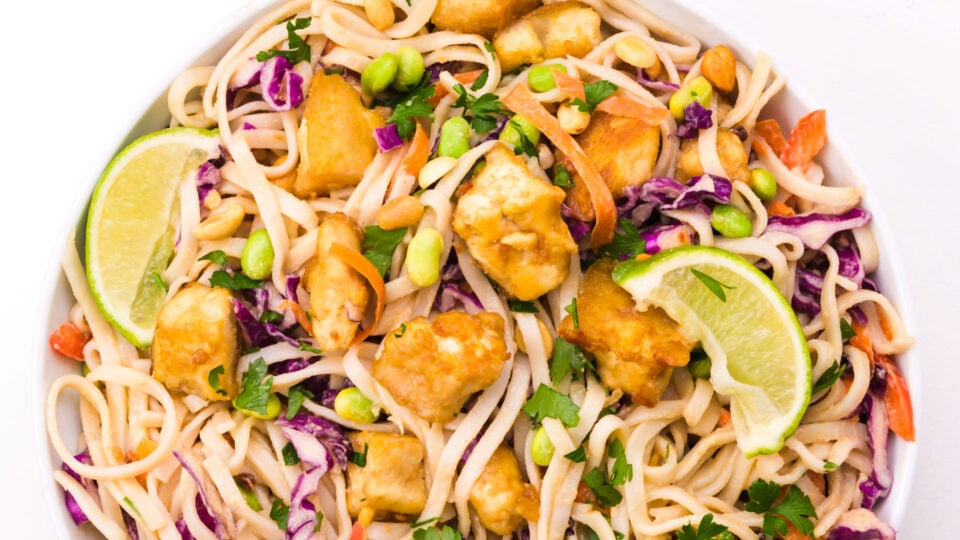 Looking down on a bowl of peanut noodle salad is topped with fried tofu and a lime slice.