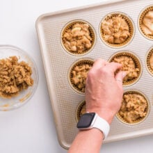 Crumble topping is being added to batter in muffin compartments.
