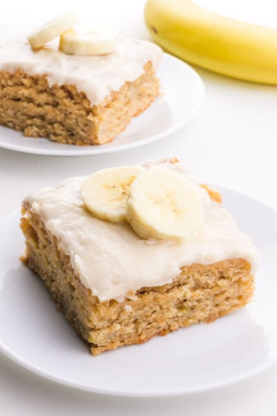 A slice of banana snack cake on a plate is topped with frosting and sliced bananas. There is another slice and a banana behind it.
