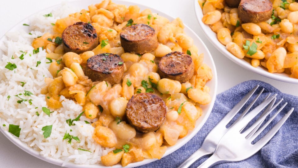Two bowls of white beans and rice with browned sausage on top sitting on a blue kitchen towel next to forks.