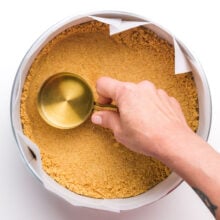 A measuring cup is being used to press graham cracker crust into a pan.