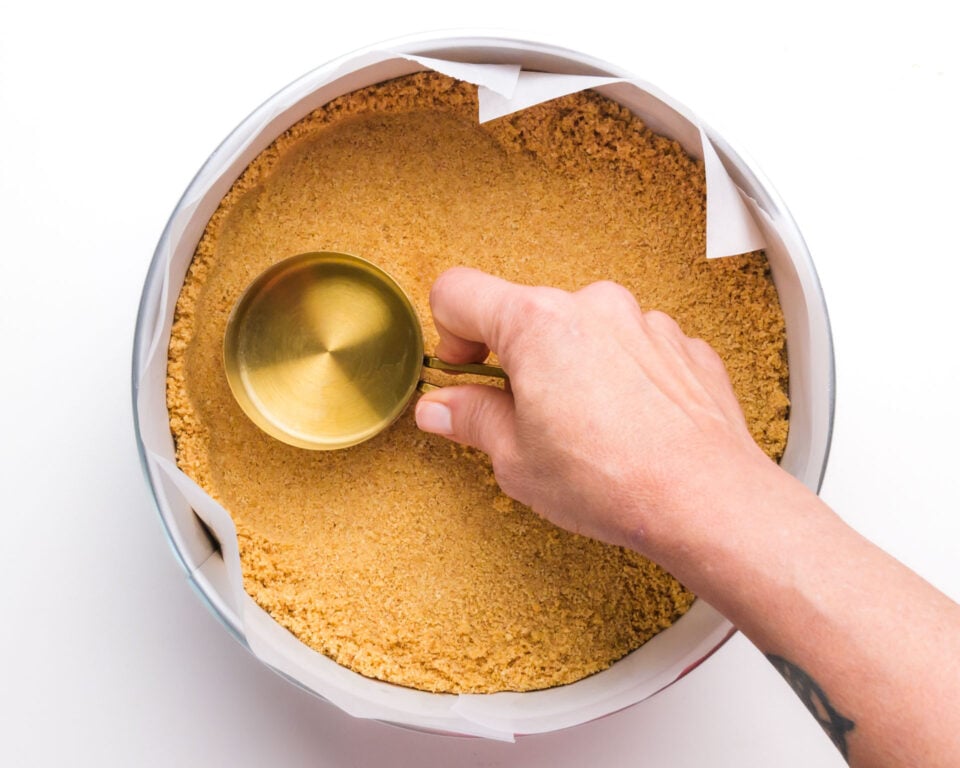 A measuring cup is being used to press graham cracker crust into a pan.