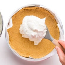 A cheesecake mixture is being spooned over graham cracker crust in a pan.