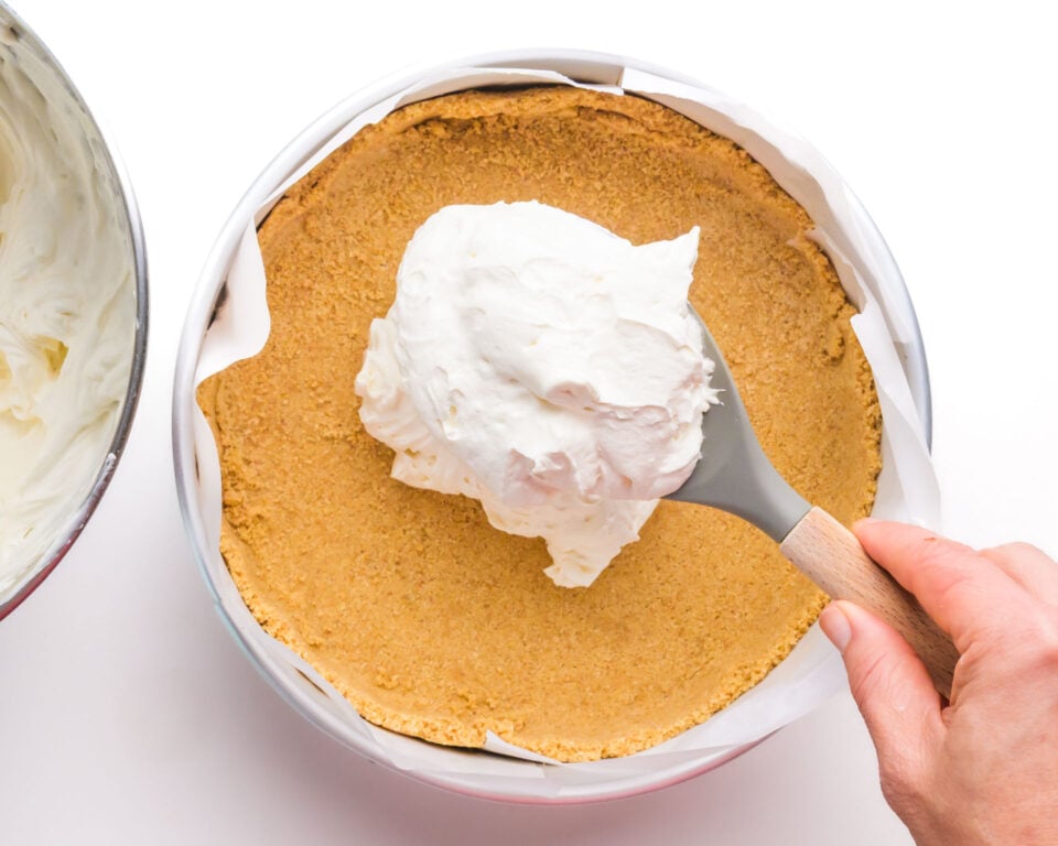 A cheesecake mixture is being spooned over graham cracker crust in a pan.