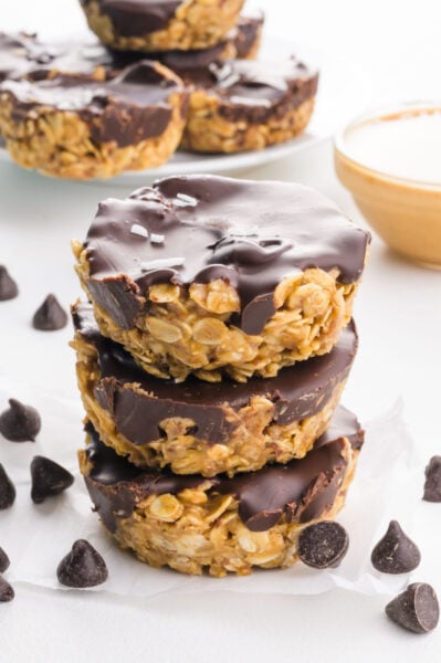 A stack of peanut butter oat cups is surrounded by chocolate chips and more of the treats in the background.