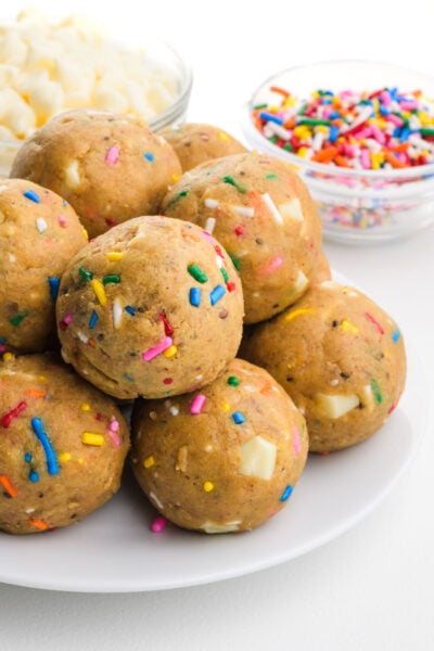 Colorful dough balls topped with sprinkles, served on a white plate, with bowls of white chocolate chips and mixed sprinkles in the background.