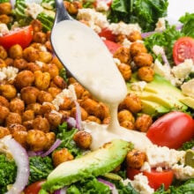 Creamy dressing is being drizzled over a kale salad with chickpeas, avocado slices, and cherry tomatoes.