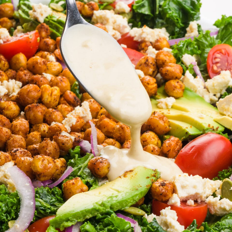Creamy dressing is being drizzled over a kale salad with chickpeas, avocado slices, and cherry tomatoes.