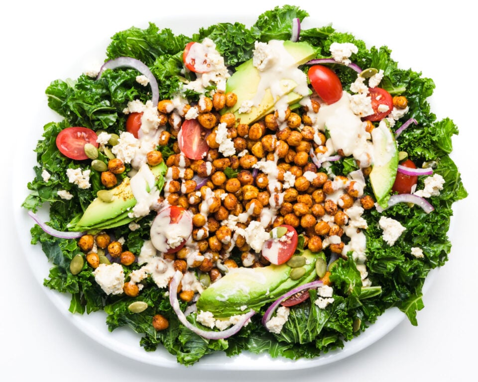 A platter holds kale salad with roasted chickpeas on top, along with sliced avocado, cherry tomatoes, and more.