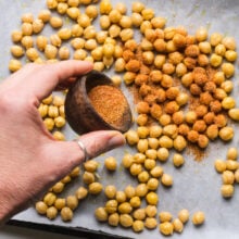 Spices are being poured over chickpeas in a pan lined with parchment paper.